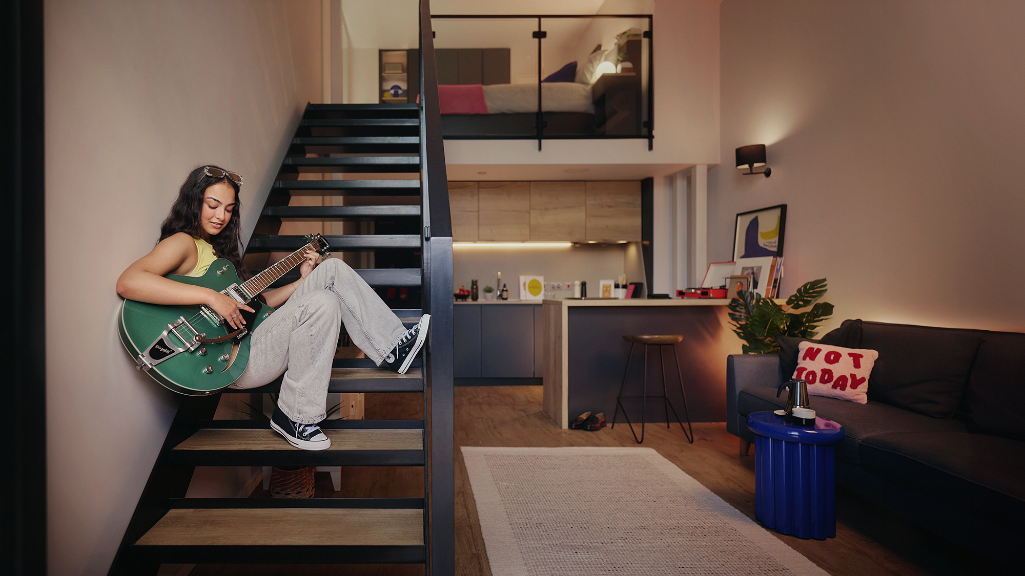 A woman sits on stairs playing a green electric guitar in a modern loft apartment. The space features a kitchen, a couch with a "Not Today" pillow, plants, and a rug. A bed is visible on the upper level.